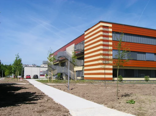 1-Klinikum-Ansbach-Erweiterungsbau-scaled-webp Ein modernes Klinikumgebäude mit rot-beiger Fassade, großen Fenstern und Außentreppen steht auf einem frisch angelegten Gelände mit jungen Bäumen
