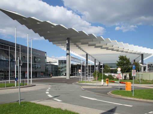 Messe_Nuernberg_4 Modernes Eingangsgebäude mit auffälligem weißen Vordach, Glasfassade und Verkehrszeichen auf einer Zufahrtsstraße unter einem teils bewölkten Himmel