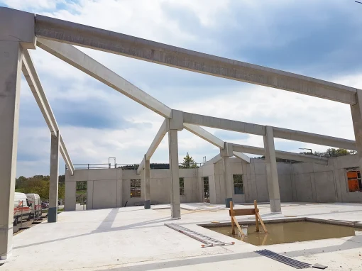 20170425_141358_web-webp Ein Rohbau mit massiven Betonstützen und Trägern steht unter bewölktem Himmel. Auf dem Boden befinden sich eine Wasserfläche, Leitern und Baumaterial