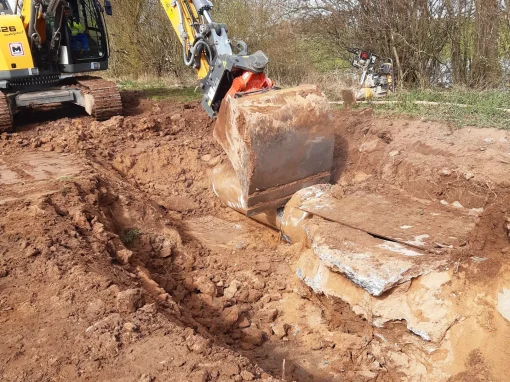 Abwasseranlage-Duerrnbuch_5-webp Ein gelber Kettenbagger steht auf einer offenen Baustelle mit lockerem Erdreich und arbeitet an einer freigelegten Betonplatte im Boden