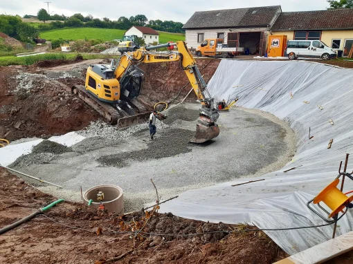 Abwasserbeseitigung-–-Markt-Taschendorf_2-1-webp Ein Bagger verteilt Schotter in einer großen Baugrube mit Folienauskleidung, ein Bauarbeiter kontrolliert die Arbeit, umgeben von landwirtschaftlichen Gebäuden und Fahrzeugen