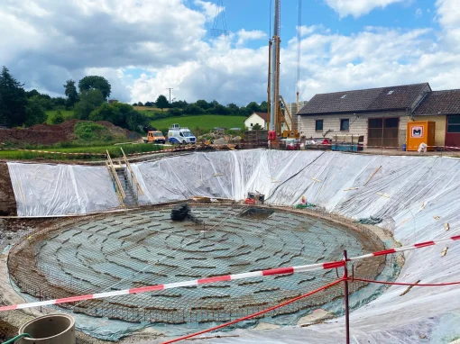 Abwasserbeseitigung-–-Markt-Taschendorf_3-webp Große Baugrube mit Metallgitter als Bewehrung auf Folie, umgeben von Absperrband und Baugeräten vor landwirtschaftlichen Gebäuden und grünem Hügel