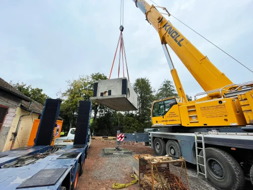 Abwasserbeseitigung-–-Markt-Taschendorf_5-webp Gelber Autokran hebt großes Betonteil mit Seilen über einer Baustelle mit Gebäuden und Fahrzeugen an einem bewölkten Tag