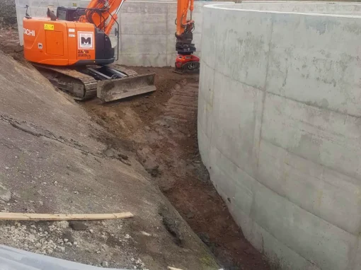 Klaeranlage-Sugenheim_4-webp Ein orangefarbener Bagger arbeitet zwischen zwei großen runden Betonbehältern auf einer Baustelle mit Erdarbeiten im ländlichen Gebiet bei bewölktem Himmel