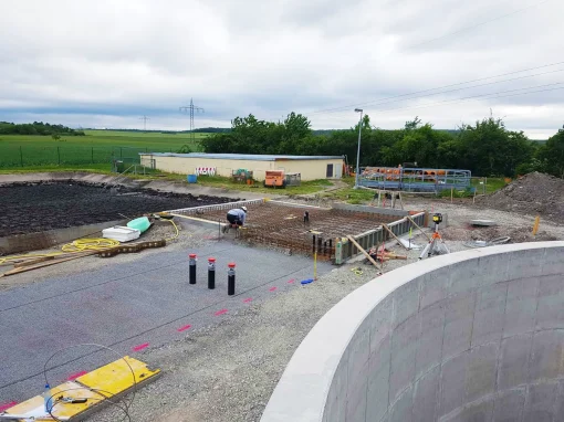 Klaeranlage-Sugenheim_5-webp Baustelle einer Kläranlage mit Bewehrung und Schalung im Vordergrund, einem Arbeiter bei Vermessungsarbeiten und Gebäuden sowie Feldern im Hintergrund