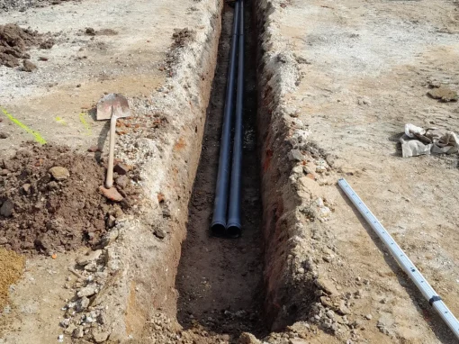 1_Kanalsanierung Hofgarten Asnabch Zwei Kunststoffrohre liegen nebeneinander in einem schmalen, frisch ausgehobenen Graben auf einer Baustelle vor einem Gebäude mit hellen Fassaden und Fenstern