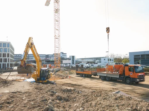 1_Tucher Campus Marienberg BA4 Baustelle mit Bagger, LKW, Kran und umliegenden Bürogebäuden. Erdarbeiten sind im Gange, verschiedene Maschinen und Materialcontainer sind im Einsatz