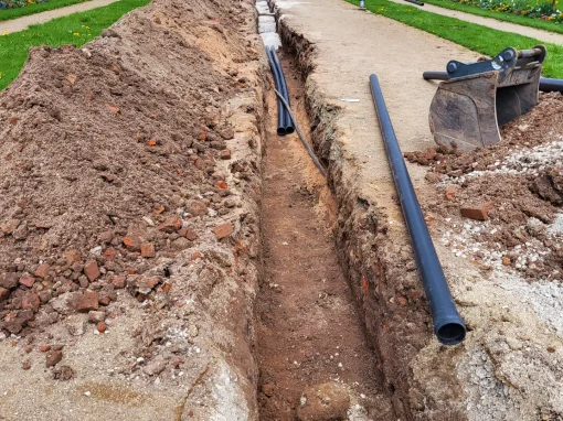 2_Kanalsanierung Hofgarten Ansbach Ein langer offener Graben mit verlegten Kunststoffrohren verläuft durch eine Gartenanlage vor einem Gebäude mit großen Rundbogenfenstern, Arbeiter sind im Hintergrund zu sehen