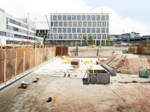 2_Tucher Campus Marienberg BA4 Baustelle mit tief ausgehobener Baugrube vor einem modernen Bürogebäude. Baumaterialien, Schubkarren und Maschinen sind auf der Fläche verteilt, links hohe Spundwände