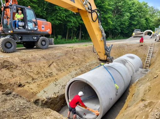2_Umbau Kläranlage Simmershofen Ein Bagger senkt ein großes Betonrohr in einen vorbereiteten Graben, Arbeiter richten es aus, im Hintergrund Maschinen, Schotter und ein Waldstück