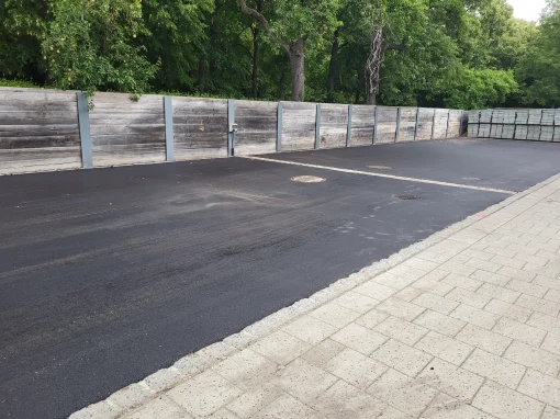 3_Kanalsanierung Hofgarten Ansabch Frisch asphaltierte Fläche mit mehreren Kanalschächten neben einem gepflasterten Gehweg, im Hintergrund eine Holzwand mit Stahlelementen und gestapelten Pflastersteinen