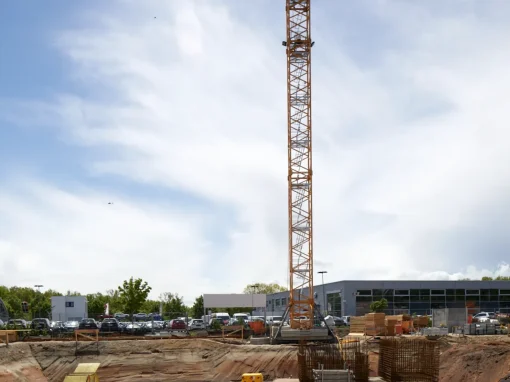 3_Tucher Campus Marienberg BA4 Großer Baukran ragt über eine Baustelle mit Fundamentarbeiten. Im Vordergrund liegen Baumaterialien und ein Erdwall, im Hintergrund parken Autos vor Bürogebäuden