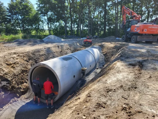 3_Umbau Kläranlage Simmershofen Zwei Arbeiter richten ein bereits verlegtes Betonrohr in einem breiten Graben aus, daneben ein Bagger und Schotterhaufen unter Bäumen bei sonnigem Wetter