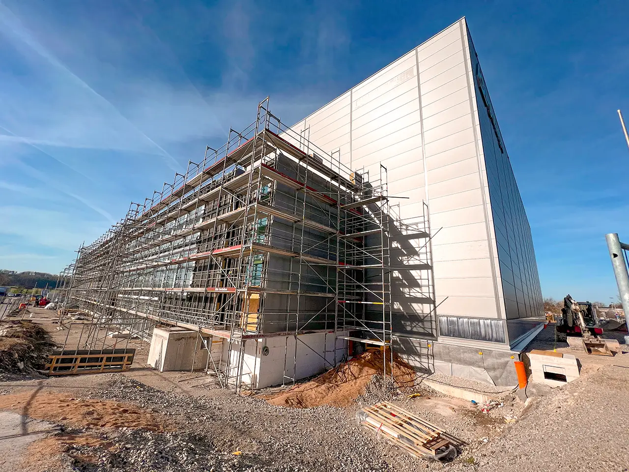 Großes Industriegebäude im Bau mit umfassendem Gerüst an der Seite unter blauem Himmel auf einem weiträumigen Baustellengelände mit Bauschutt und Maschinen