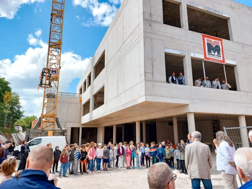 5_Grundschule Schalkhausen Viele Kinder und Erwachsene stehen vor einem Rohbau einer Grundschule. Mehrere Personen schauen aus Fenstern im Obergeschoss zu. Ein Kran mit dem Schild „Moezer“ ist im Hintergrund zu sehen