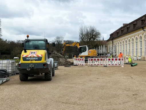 5_Kanalsanierung Hofgarten Ansabch Baustelle vor einem Gebäude mit Bauzaun, Bagger und Radlader; mehrere Bauarbeiter in Warnkleidung arbeiten im abgesperrten Bereich