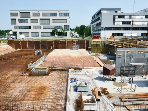 5_Tucher Campus Marienberg BA4_ (Titelbild) Baustelle mit sichtbarer Bewehrung, Schalung und ersten Betonwänden. Im Hintergrund stehen moderne Bürogebäude, vorne liegen Baumaterialien und Baugerüste