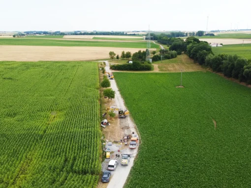 5_Umbau Kläranlage Simmershofen Luftaufnahme einer Baustelle zwischen zwei großen Feldern mit landwirtschaftlicher Nutzung, Baufahrzeuge und Material lagern entlang eines Feldwegs, im Hintergrund Windräder und Strommasten