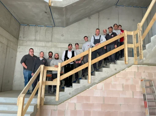 6_Grundschule Schalkhausen Eine Gruppe von Handwerkern steht lächelnd auf einer Treppe in einem Rohbau, im Vordergrund ist ein Holzgeländer, im Hintergrund Wände aus Beton und Stein