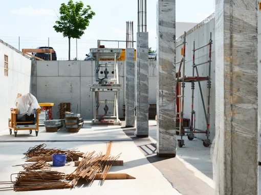 6_Tucher Campus Marienberg BA4 Baustellenbereich mit fertiggestellten Betonwänden und verpackten Stützen. Im Vordergrund liegen Bewehrungseisen und Baumaterialien, ein Baum ragt über die Mauer hinaus