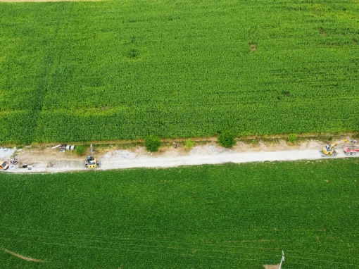 6_Umbau Kläranlage Simmershofen Luftaufnahme eines Feldwegs mit mehreren Fahrzeugen und Maschinen, die sich entlang eines langen, schmalen Streifens zwischen dicht bewachsenen grünen Feldern verteilen