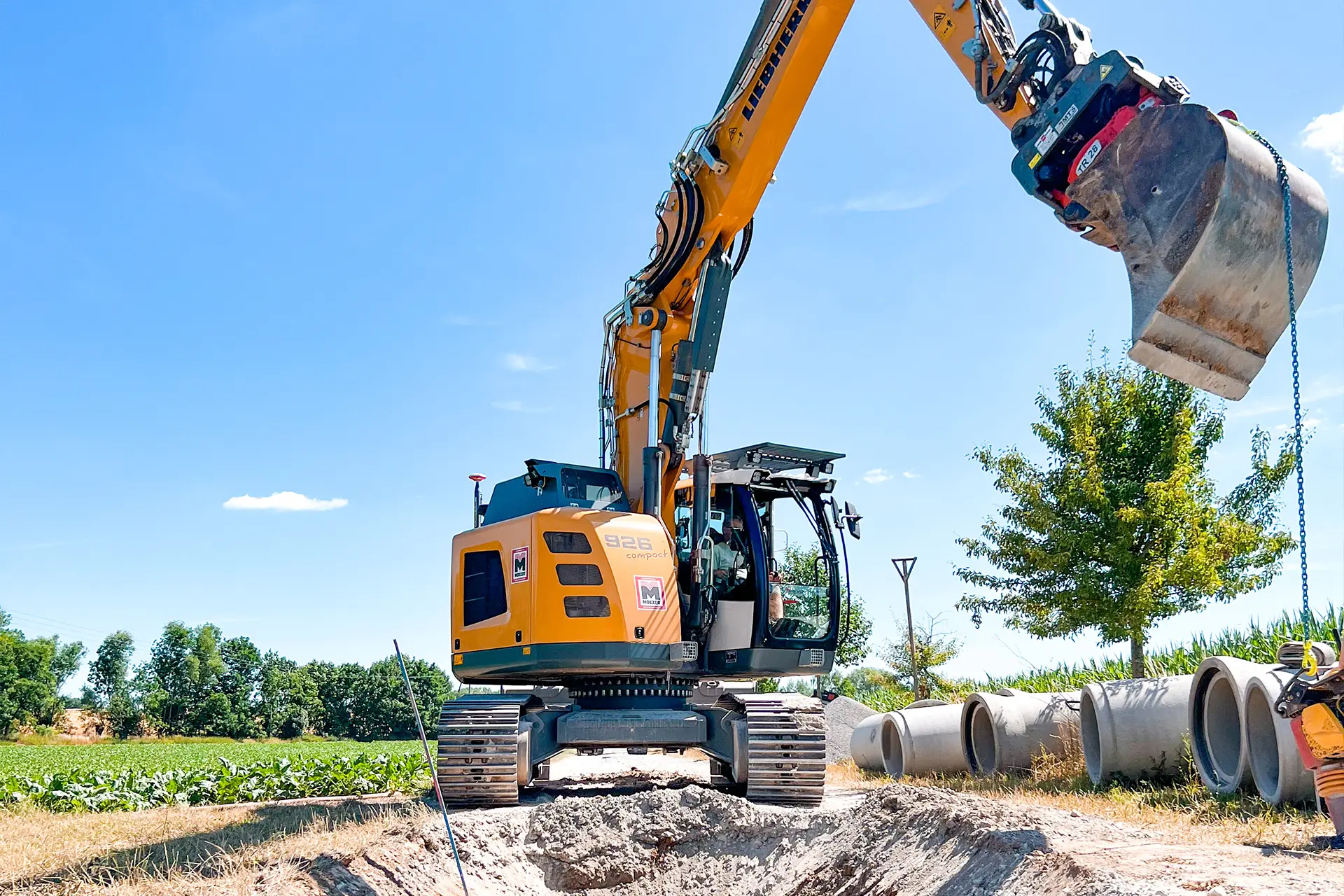 bagger-maschinenpark-webp Ein gelber Kettenbagger mit Anbaugerät kippt Erde auf eine Baustelle, während im Hintergrund Bäume und blauer Himmel mit Wolken zu sehen sind